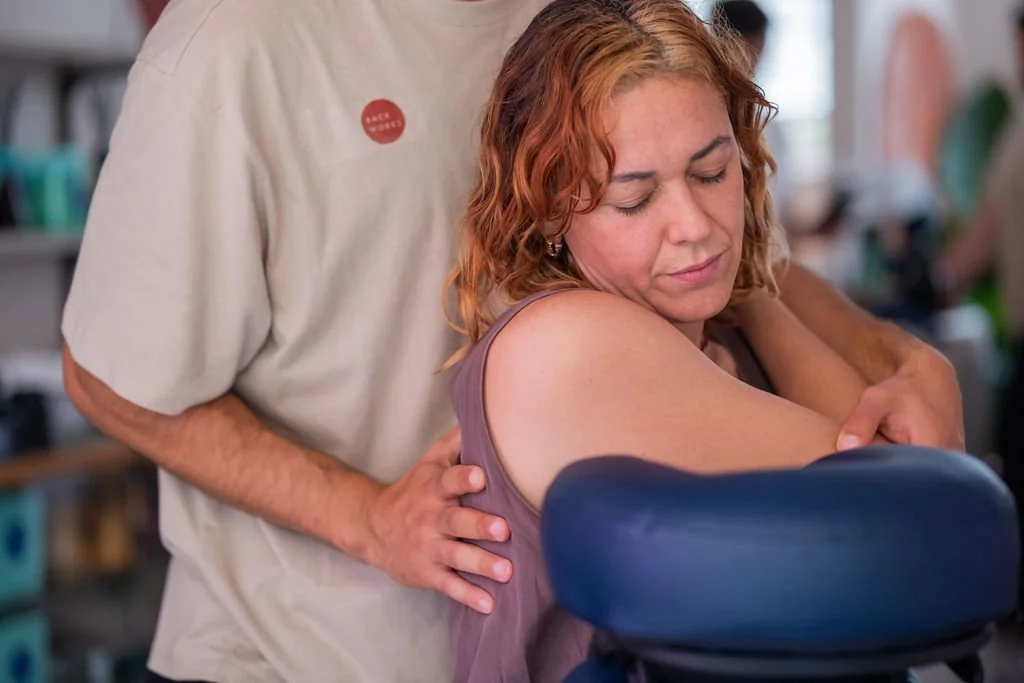 A woman receiving a shoulder massage from a man in a food service uniform in a busy environment.