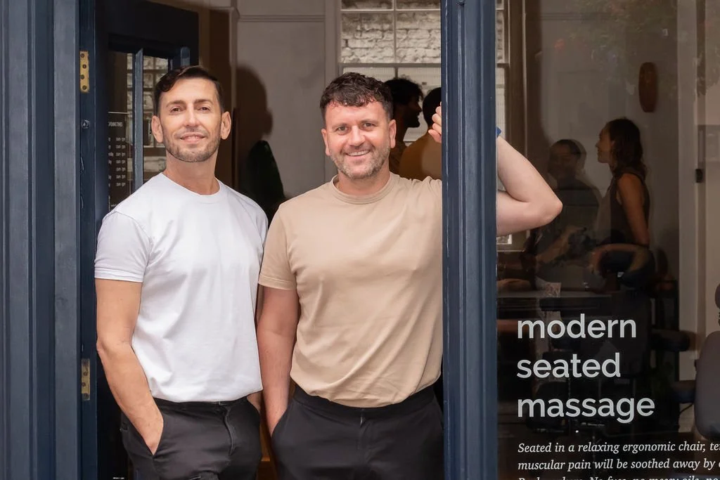 Two men standing in a doorway, smiling, with a glass door reflecting three women inside a massage spa. Text on the door reads 'modern seated massage'.