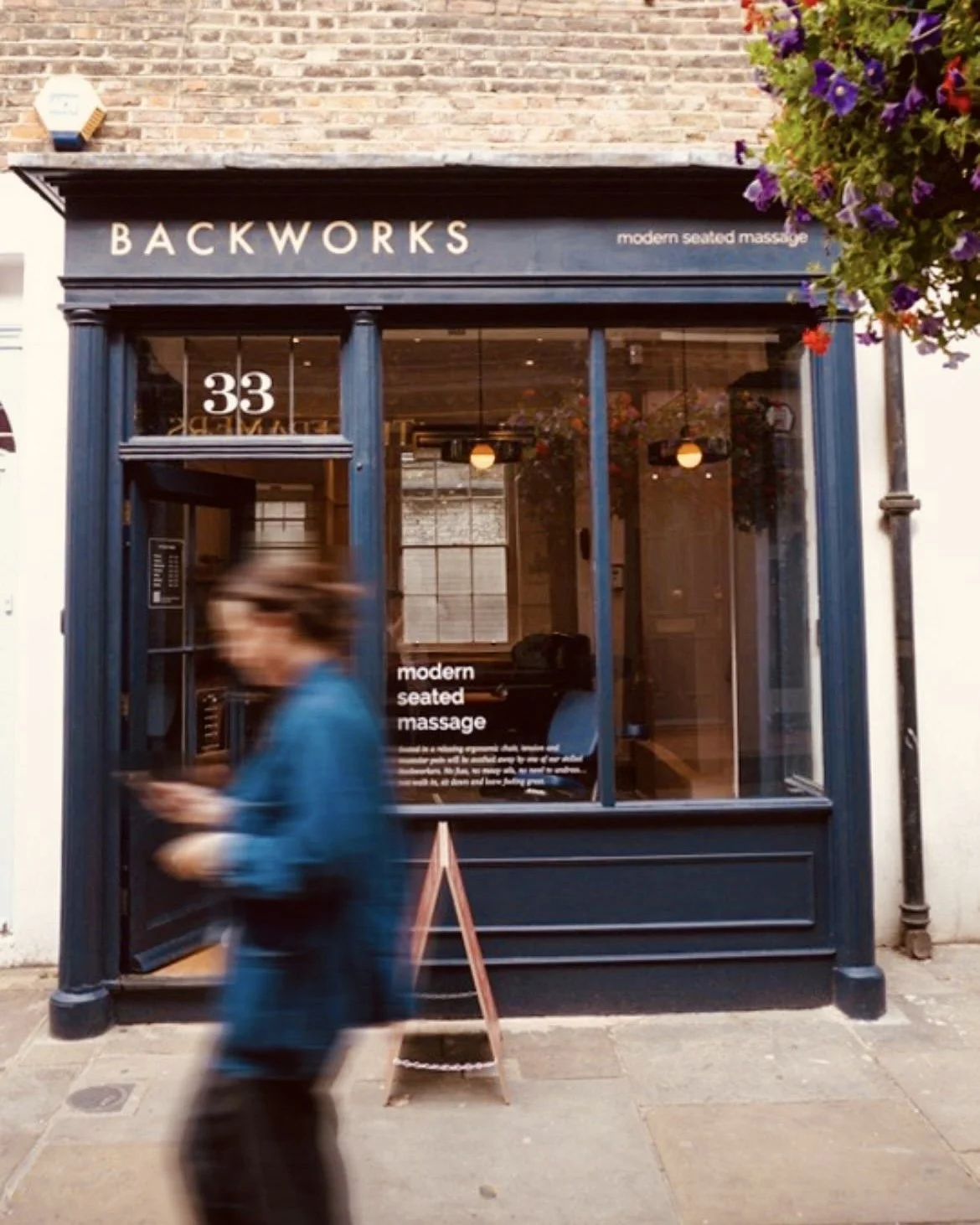 The storefront of Backworks, a modern seated massage business at 33, with large glass windows and a black facade. A person in a blue jacket is walking past and appears blurred due to motion.