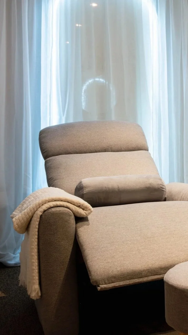 A beige upholstered armchair with a matching cushion and blanket draped over the armrest, positioned in front of sheer white curtains.