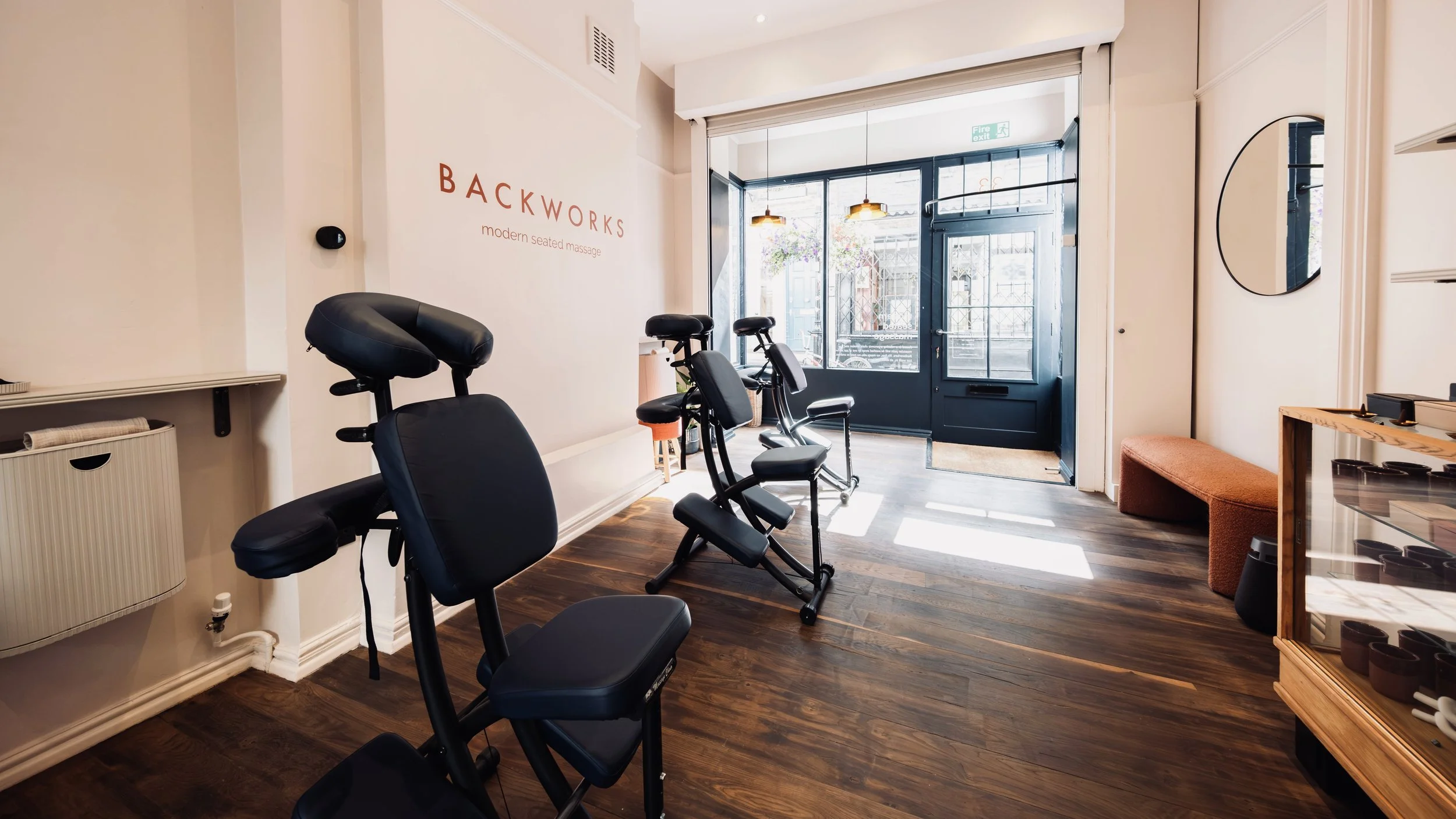 Interior of a massage studio with black massage chairs lined up, a wooden bench, and a glass door leading outside. The walls are painted light, with a sign that reads 'BACKWORKS modern seated massage,' and there is sunlight coming through the door.