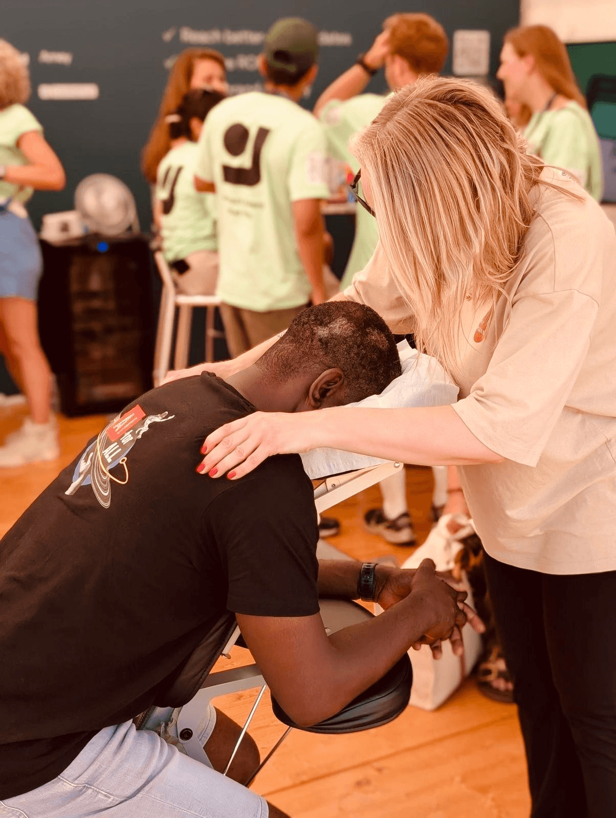 A woman with blonde hair is comforting a man sitting on a massage table, his head bowed and hands clasped. In the background, several people are standing and talking in a room with dark walls and wooden floor.
