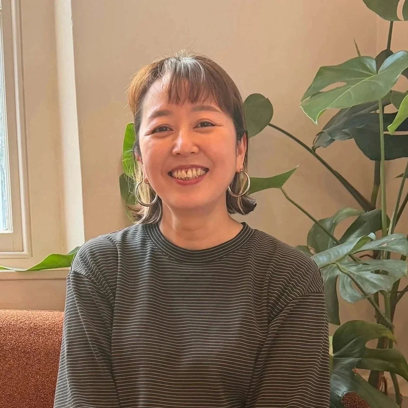Smiling woman with short brown hair and hoop earrings sitting in front of large green plants indoors.