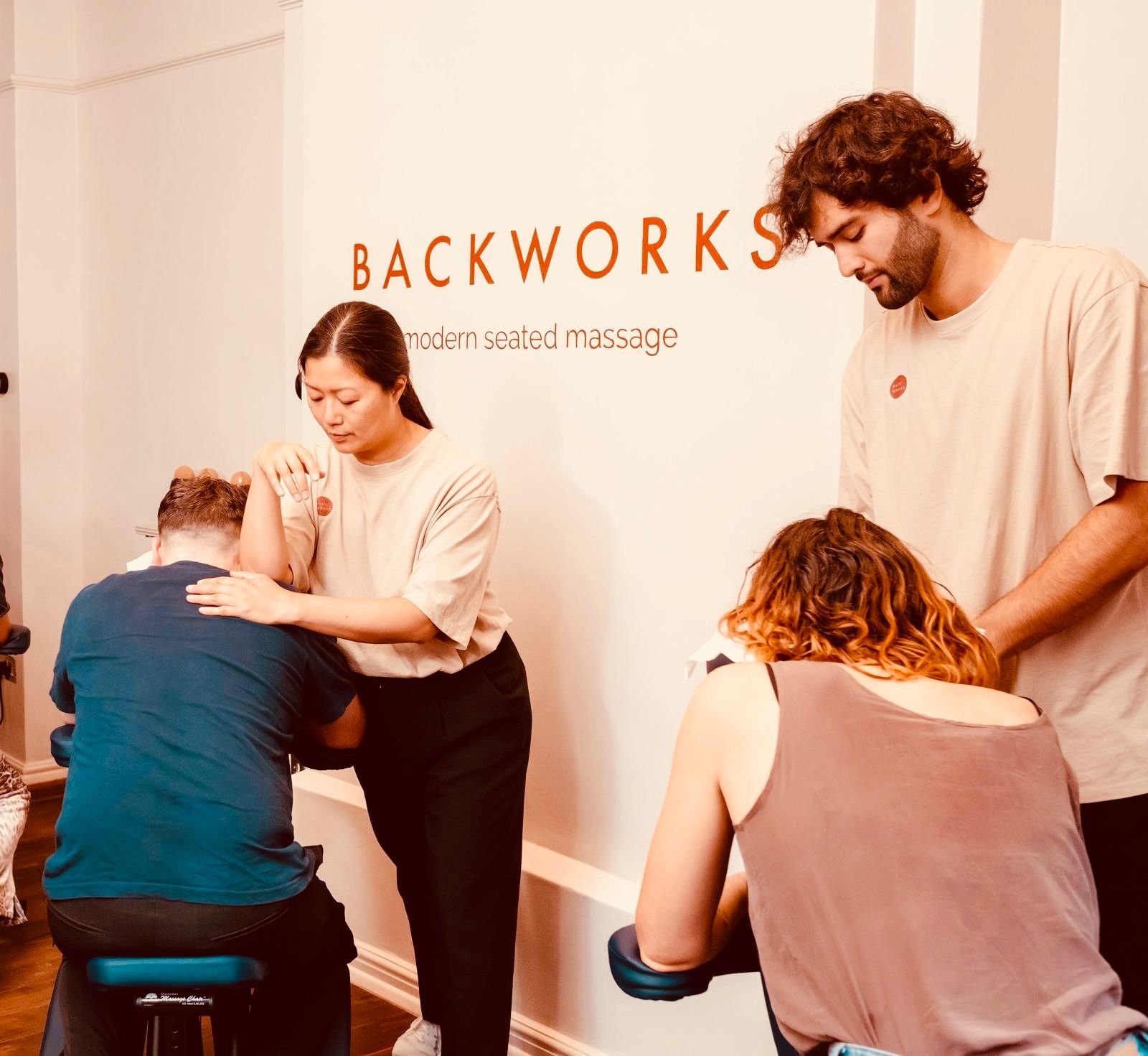 People receiving seated massage therapy at a wellness center with a sign reading 'Backworks modern seated massage' in the background.