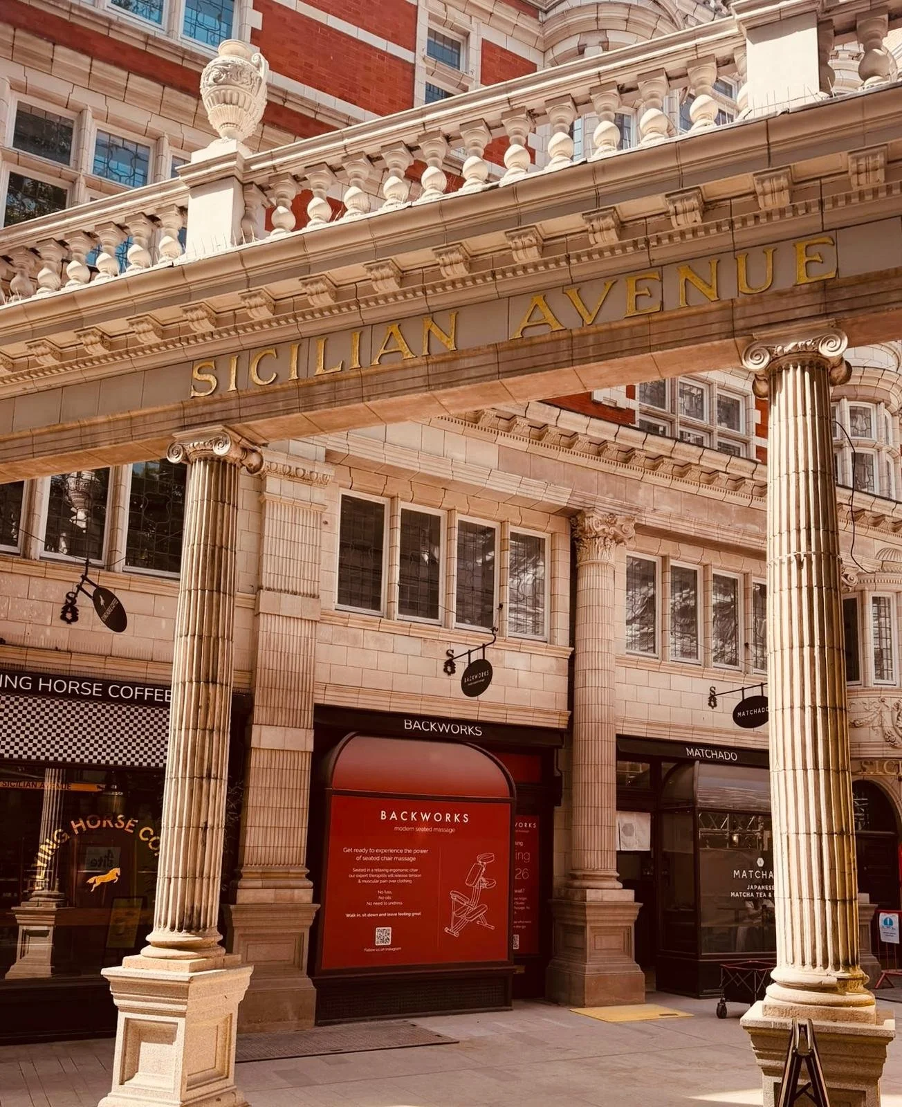 The facade of a building with classical columns and a sign that reads 'Sicilian Avenue' in gold letters. Below are shopfronts, including Backworks and other stores.