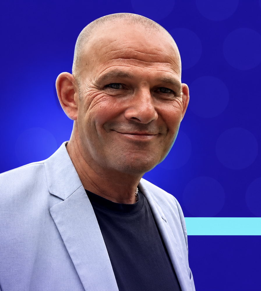 A bald man smiling, wearing a light gray jacket and a black t-shirt, standing against a blue background with circular light patterns.