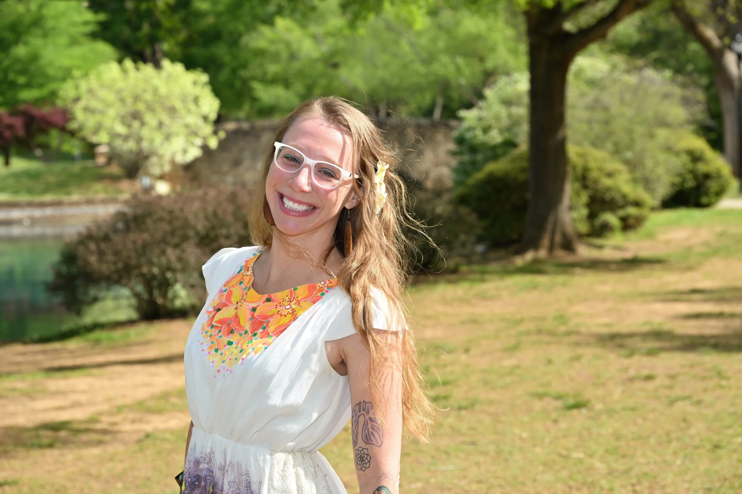 A smiling person with long wavy hair and glasses standing outdoors in a park or garden with green trees and grass.