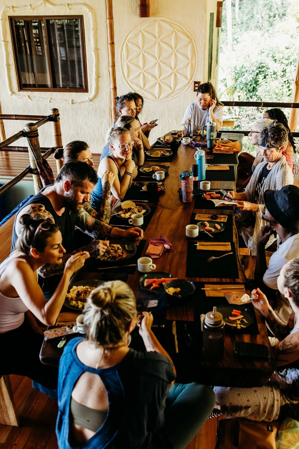 A group of people gathered around a long wooden dining table in a cozy, rustic restaurant with natural light, enjoying a meal together.