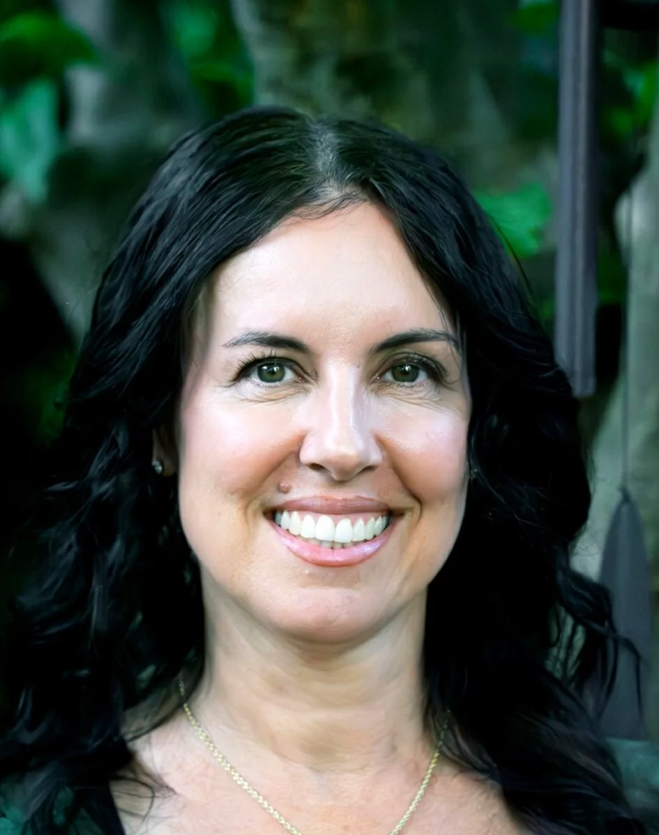 A person with black, wavy hair and green eyes smiling outdoors, with blurred green foliage in the background.