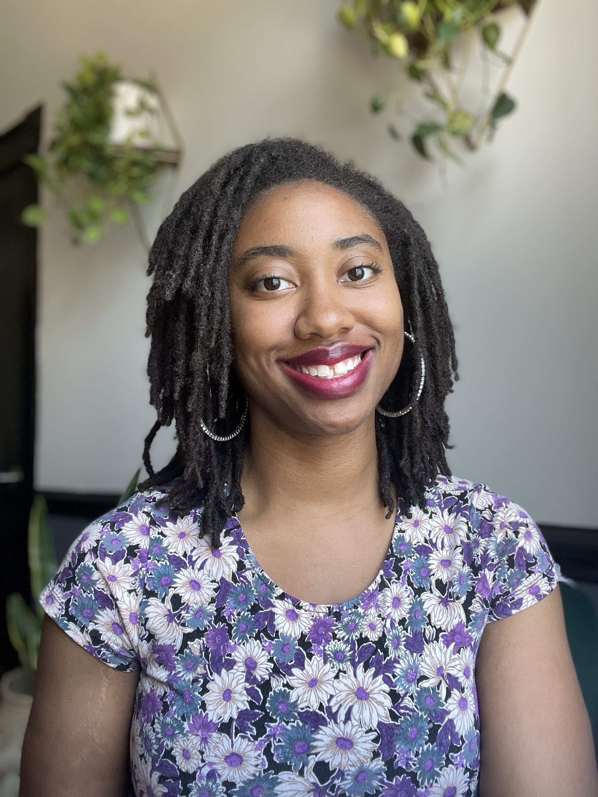 A person smiling with dark, shoulder-length locs and hoop earrings wearing a purple, white, and black floral-patterned shirt. In the background, there are two hanging plants with green leaves against a plain wall.