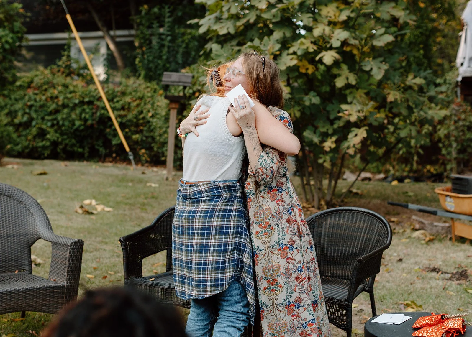 Two women hug each other outdoors, with trees in the background, one wearing a white sleeveless top and plaid skirt, and the other in a floral dress, both smiling and embracing.