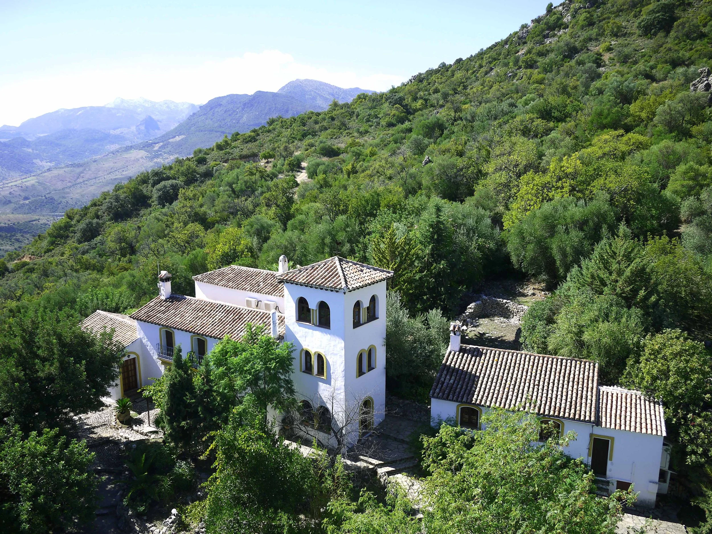A white Mediterranean-style house with clay tile roof, surrounded by lush green trees, set on a hillside with mountain range in the background.