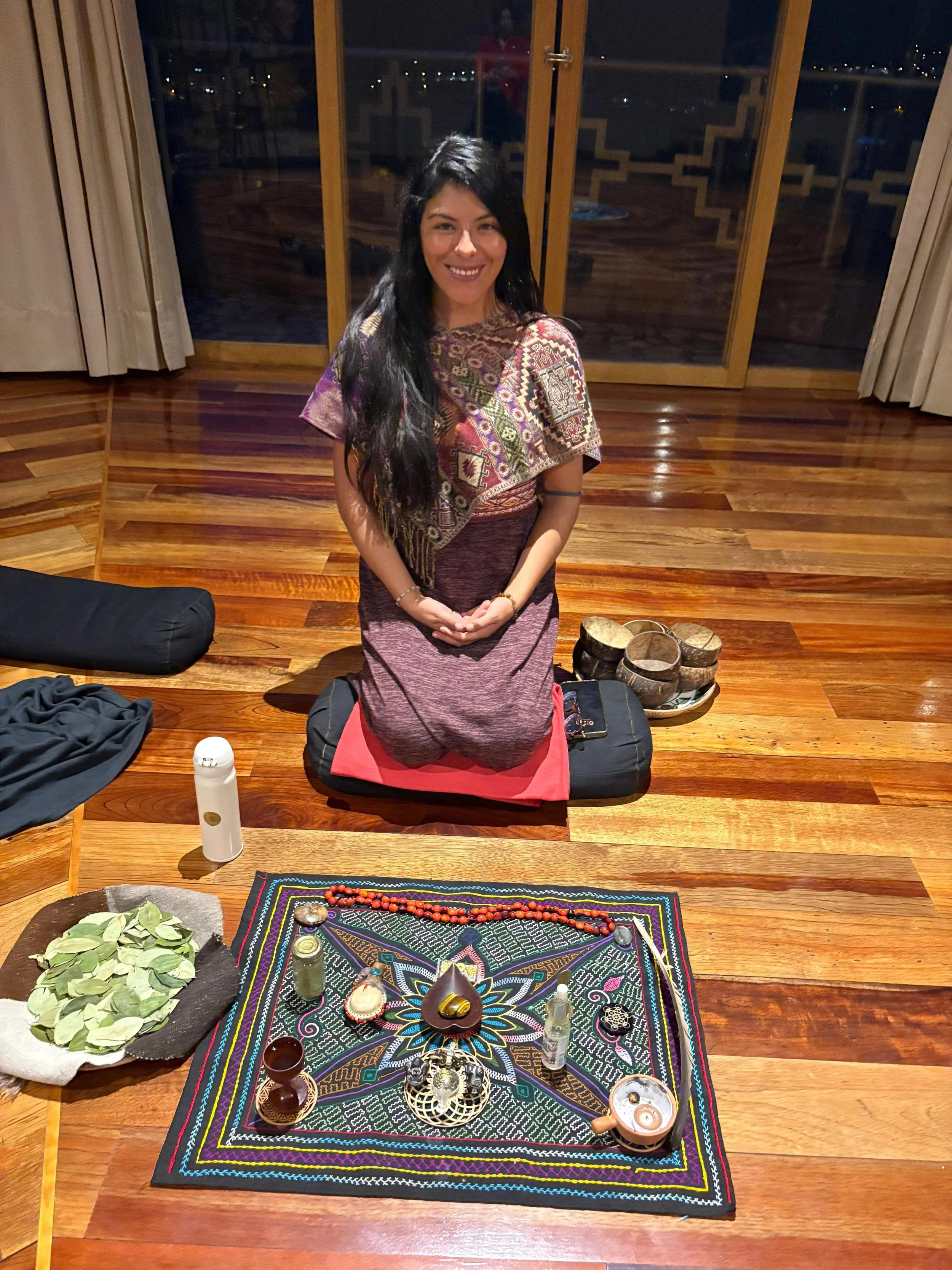 A person with long brown hair knees in front of a square cloth filled with trinkets and colors. She has a soft smile and wears traditional Peruvian clothing.