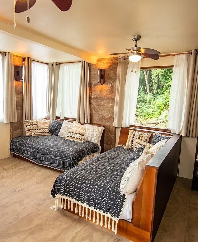 A cozy living room with two sofas covered in black and white patterned blankets, multiple pillows, and large windows with white curtains overlooking greenery.