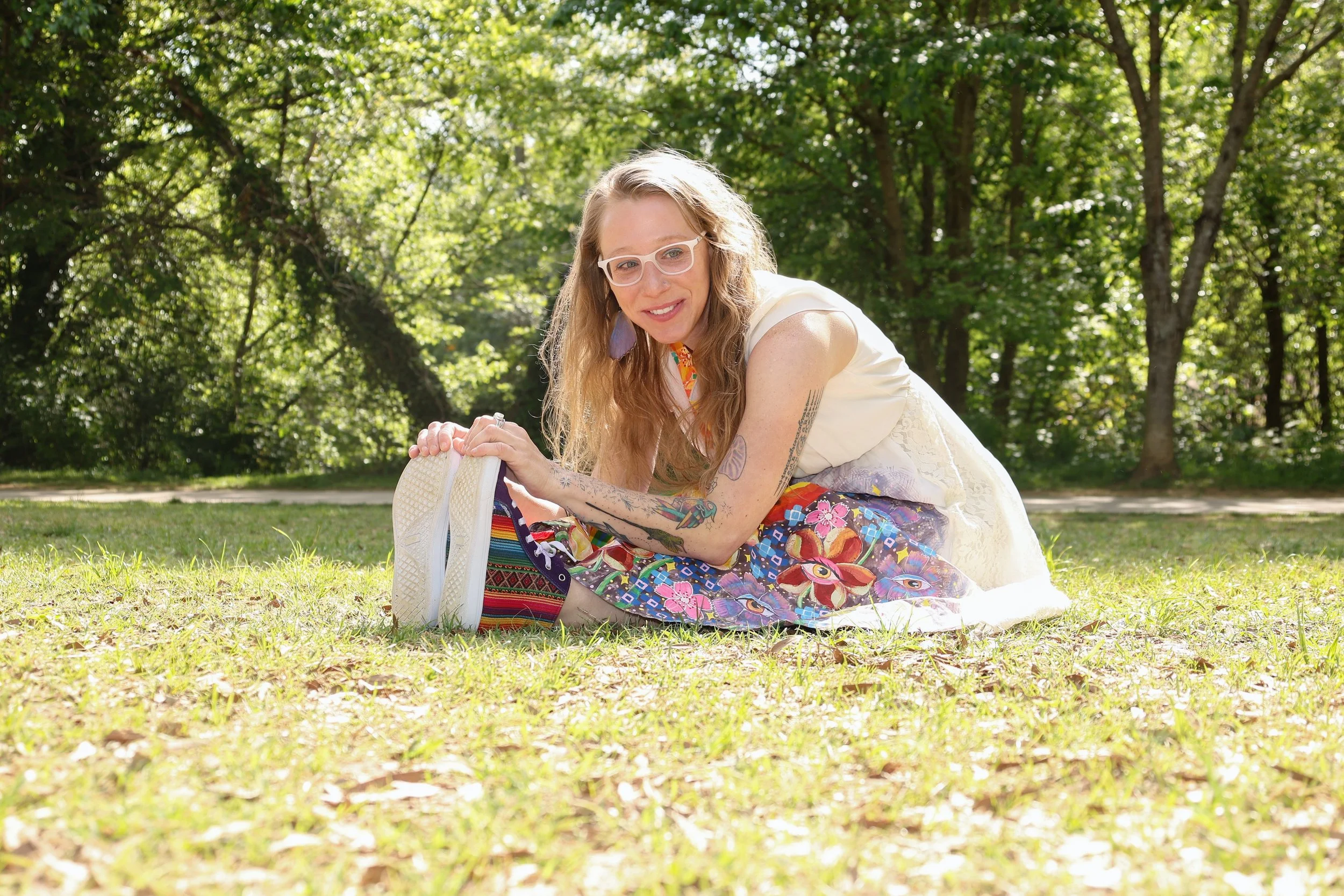 A person with curly blonde hair, glasses, and tattoos on her arms, is sitting on the grass outdoors in a wooded area with green trees and sunlight filtering through the leaves. The person is smiling and leaning forward with hands on a white bag.