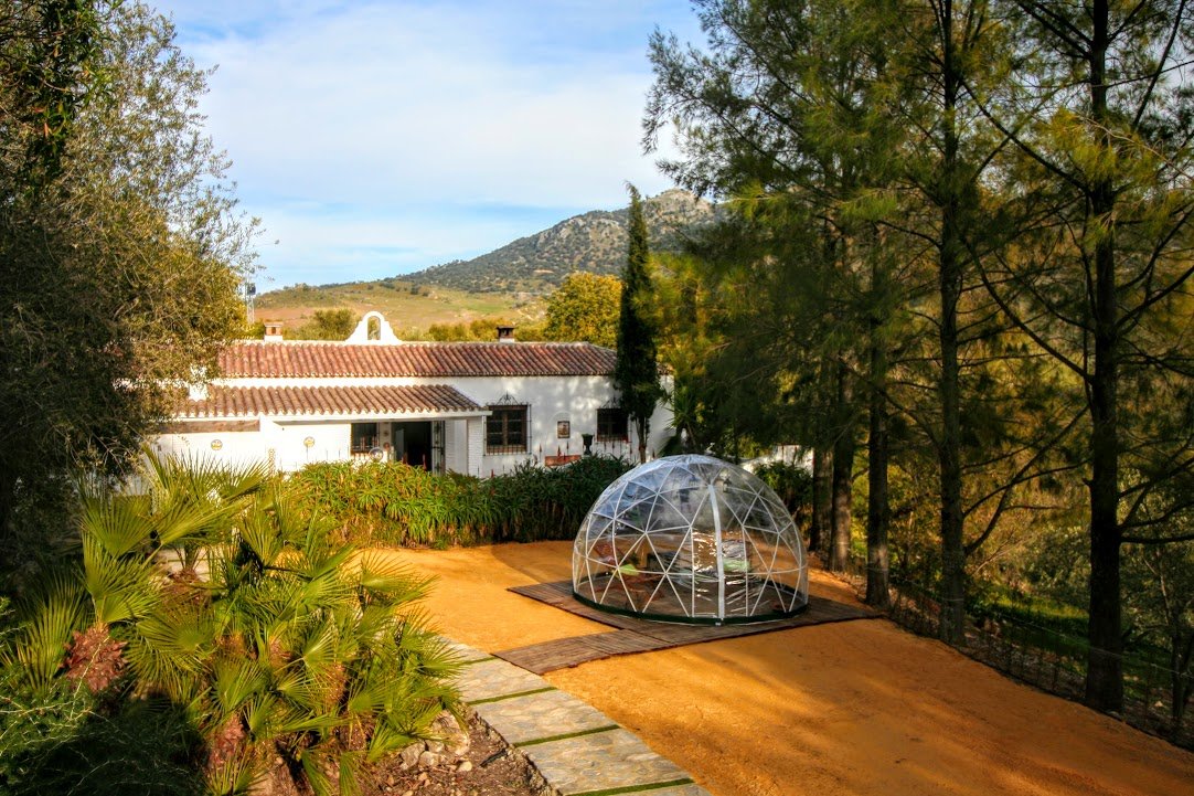 A white house with a red tile roof in a wooded area with a mountain in the background. In front of the house, there is a transparent geodesic dome structure on the ground.