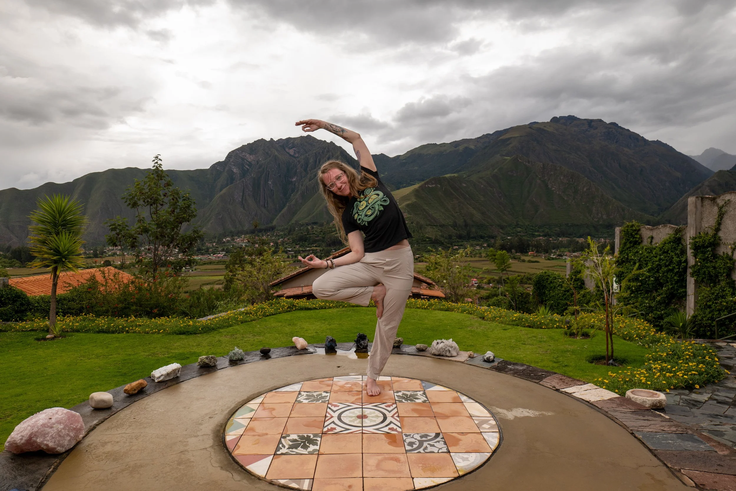 A blonde-haired person standing on one foot and balancing with backdrop of mountains with clouds.