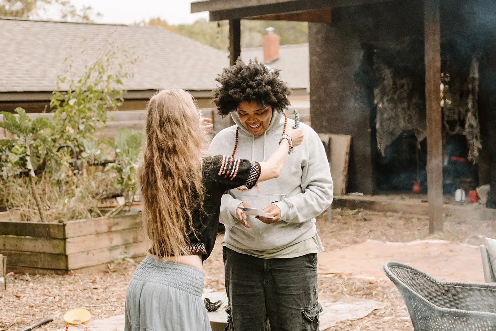 Two women standing outdoors, one with long curly hair and the other with short curly hair, smiling and engaging with each other; the woman with long hair is touching the other's necklace while the other holds a phone, with a garden and a wood structure in the background.
