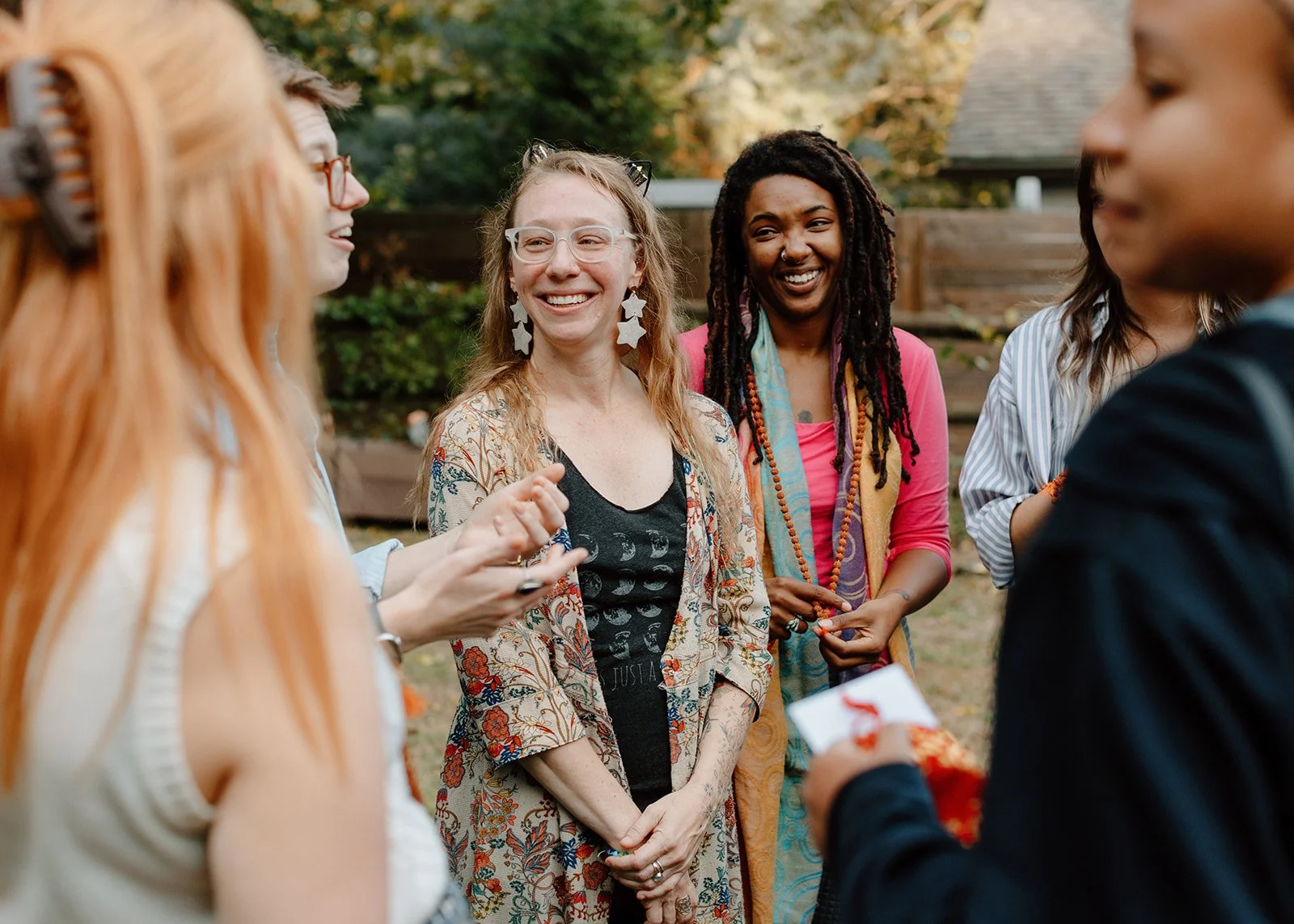 A group of five women talking and laughing outdoors during fall, with trees and a house in the background.