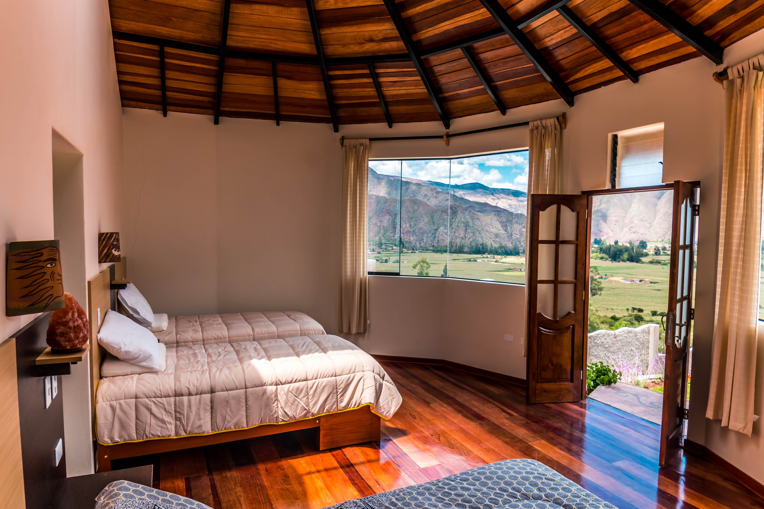 Two made beds in a circular style room that faces large windows and features an open balcony with a Mountain View and blue skies.