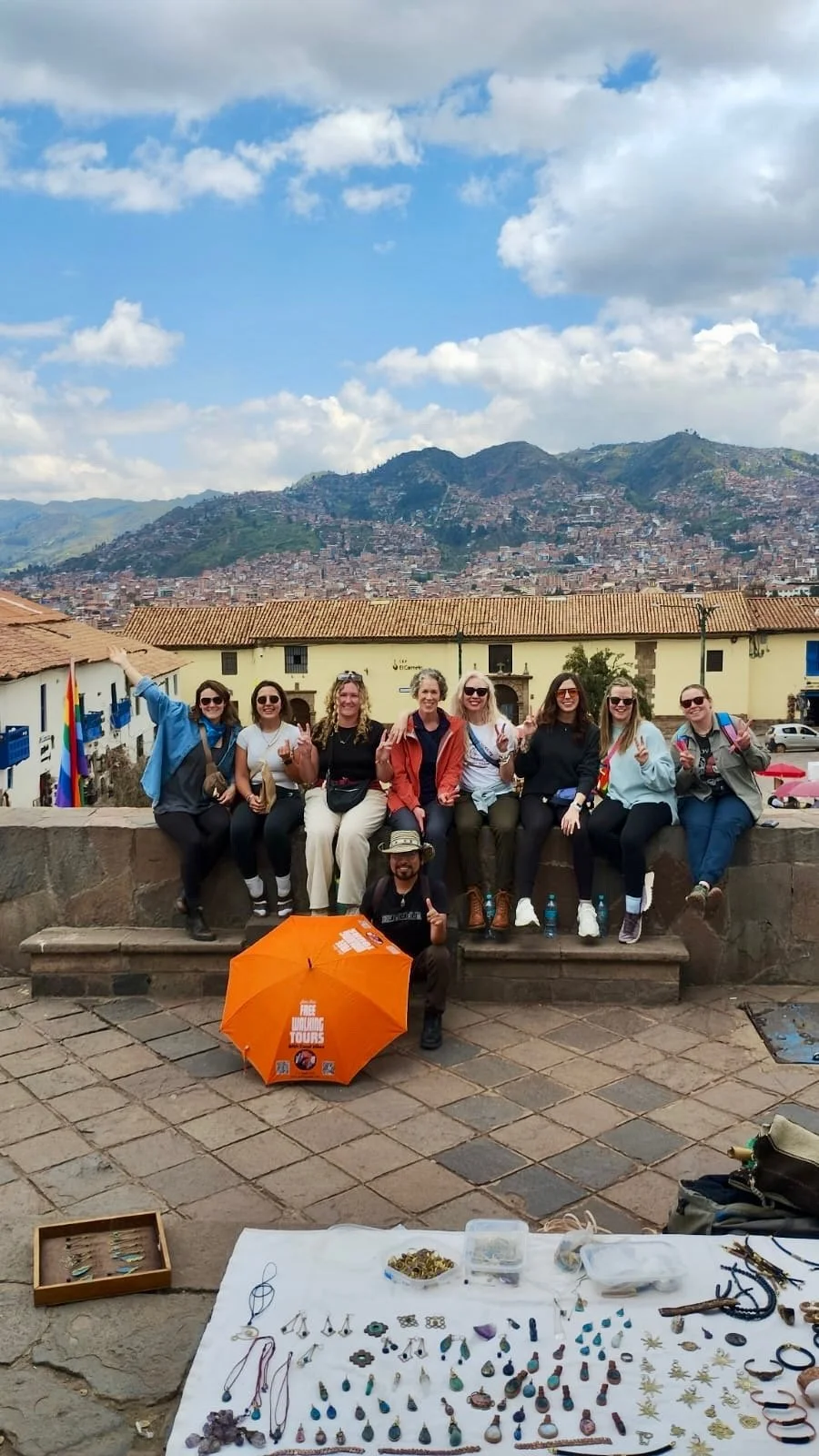 A group of 8 people sit on a ledge in a city setting with another person in front kneeling with an open orange umbrella. The background is looking down onto a city setting with mountains surrounded. Everyone is smiling.
