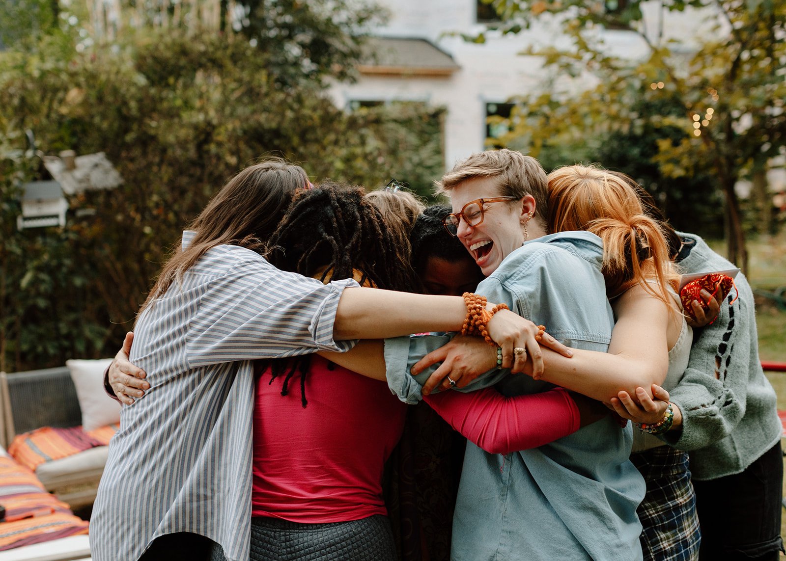 A diverse group of friends hugging and smiling outdoors in a backyard with trees and a house in the background.