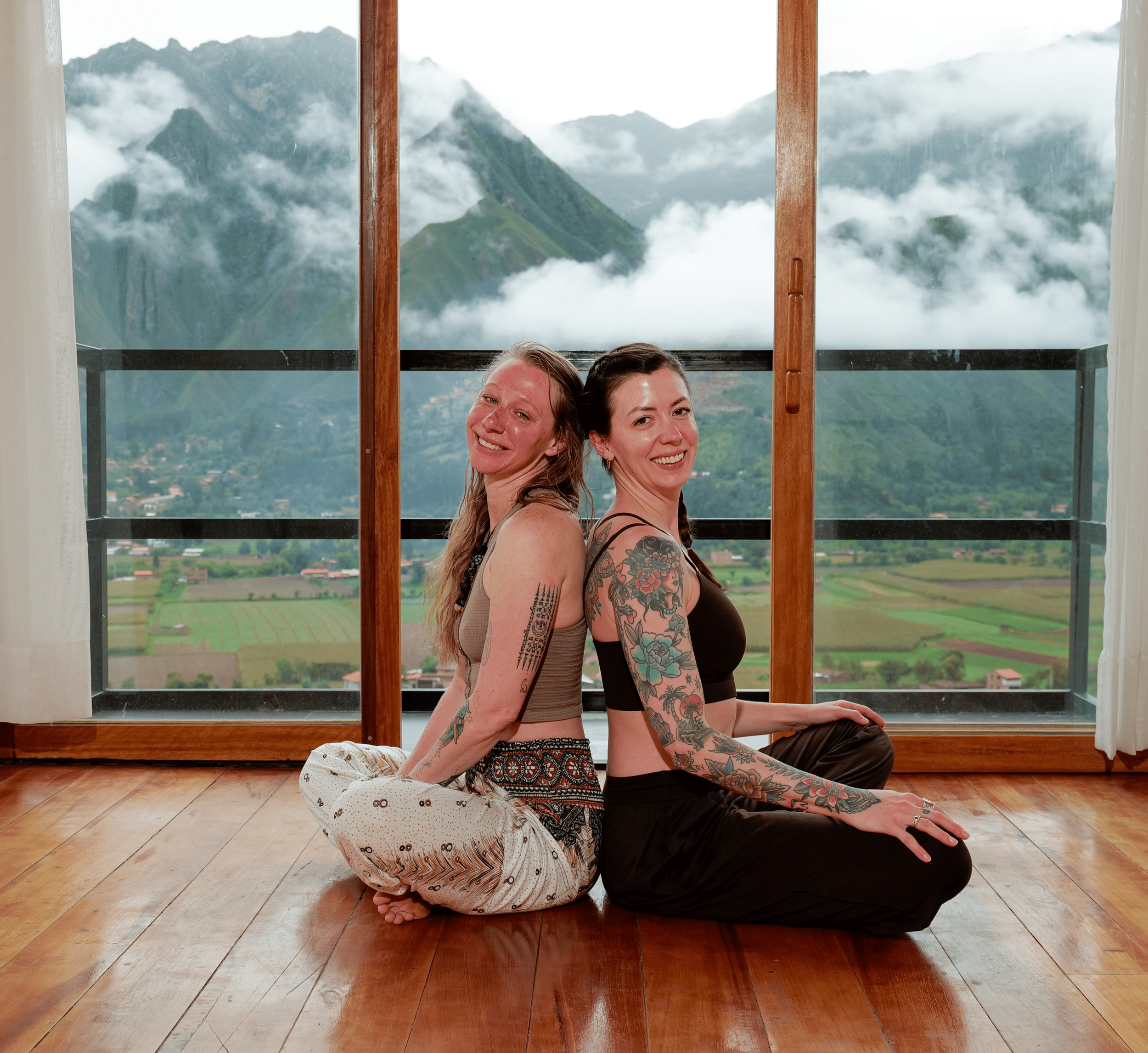 Two people sit back to back with their heads touch and they are smiling. There is a mountain backdrop with clouds.
