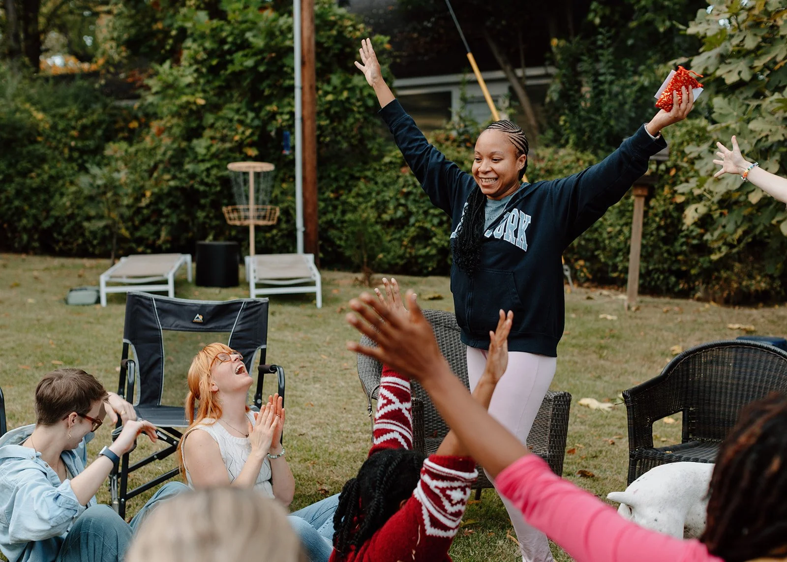 A group of friends celebrating outdoors, with a person standing with arms raised in excitement, while others sit and cheer around.