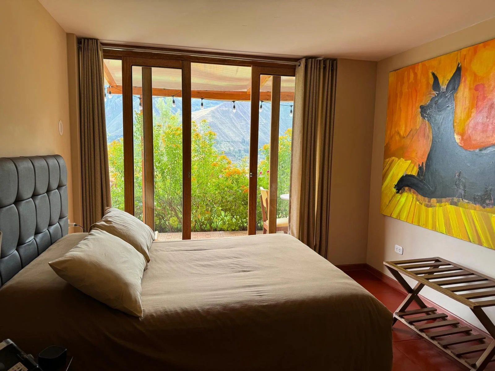 A made bed in a square room with an open door and patio behind it. The patio has tall green plants that serve as the wall with mountains in the background.