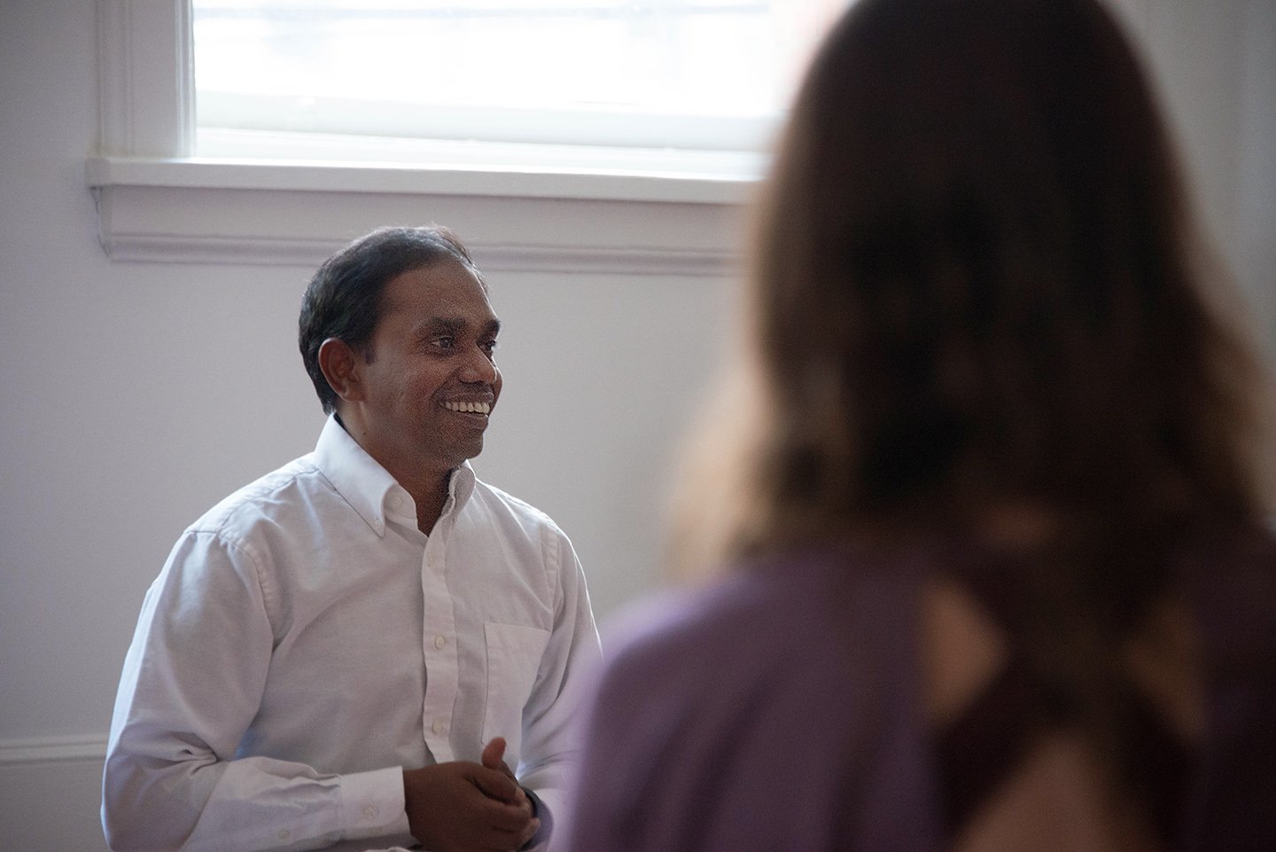A person in a white shirt smiling and talking to someone whose back is facing the camera, in a room with a window in the background.