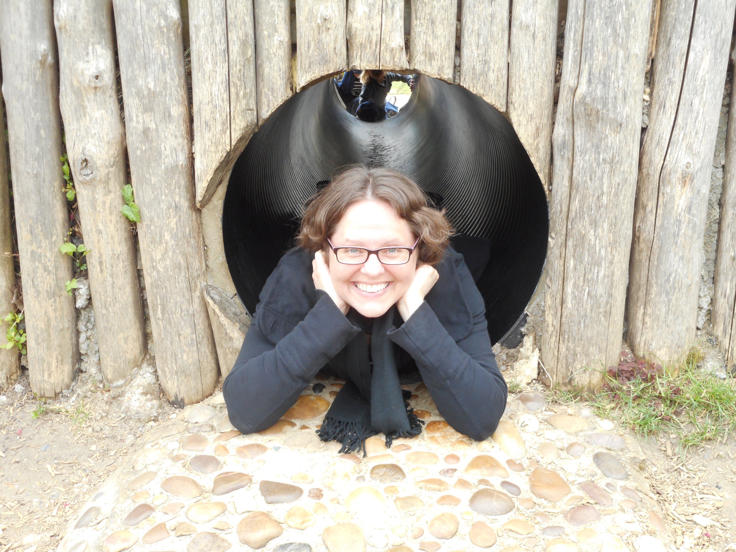 A person with glasses smiling and resting her chin on her hands, crouched at the entrance of a large pipe or tunnel built into a wooden structure, with children visible inside the tunnel.