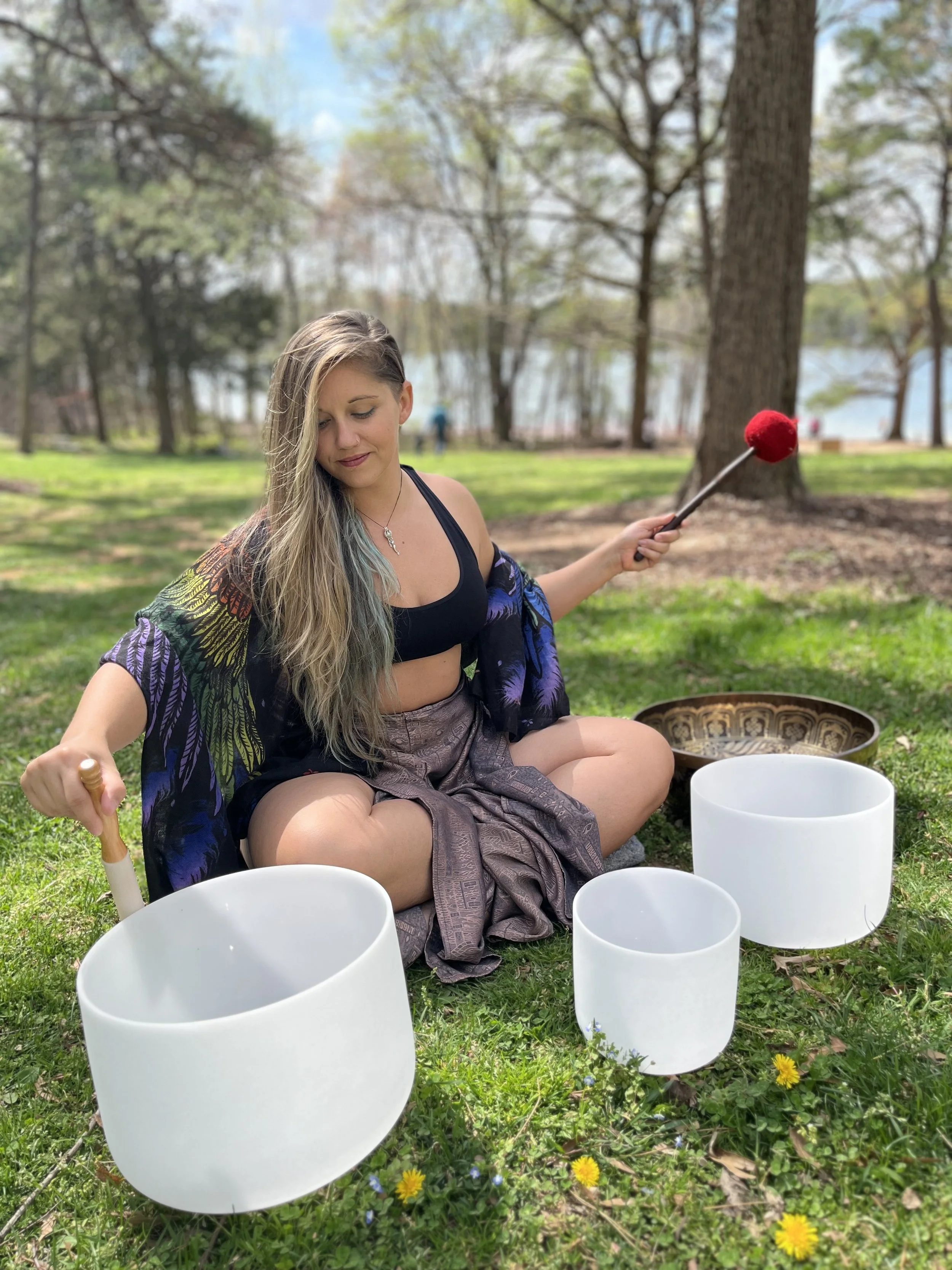 A person with long blonde hair sitting cross-legged on the grass in a park, playing crystal singing bowls surrounded by trees and overlooking a lake, holding a mallet in the right hand and looking down with a gentle expression.