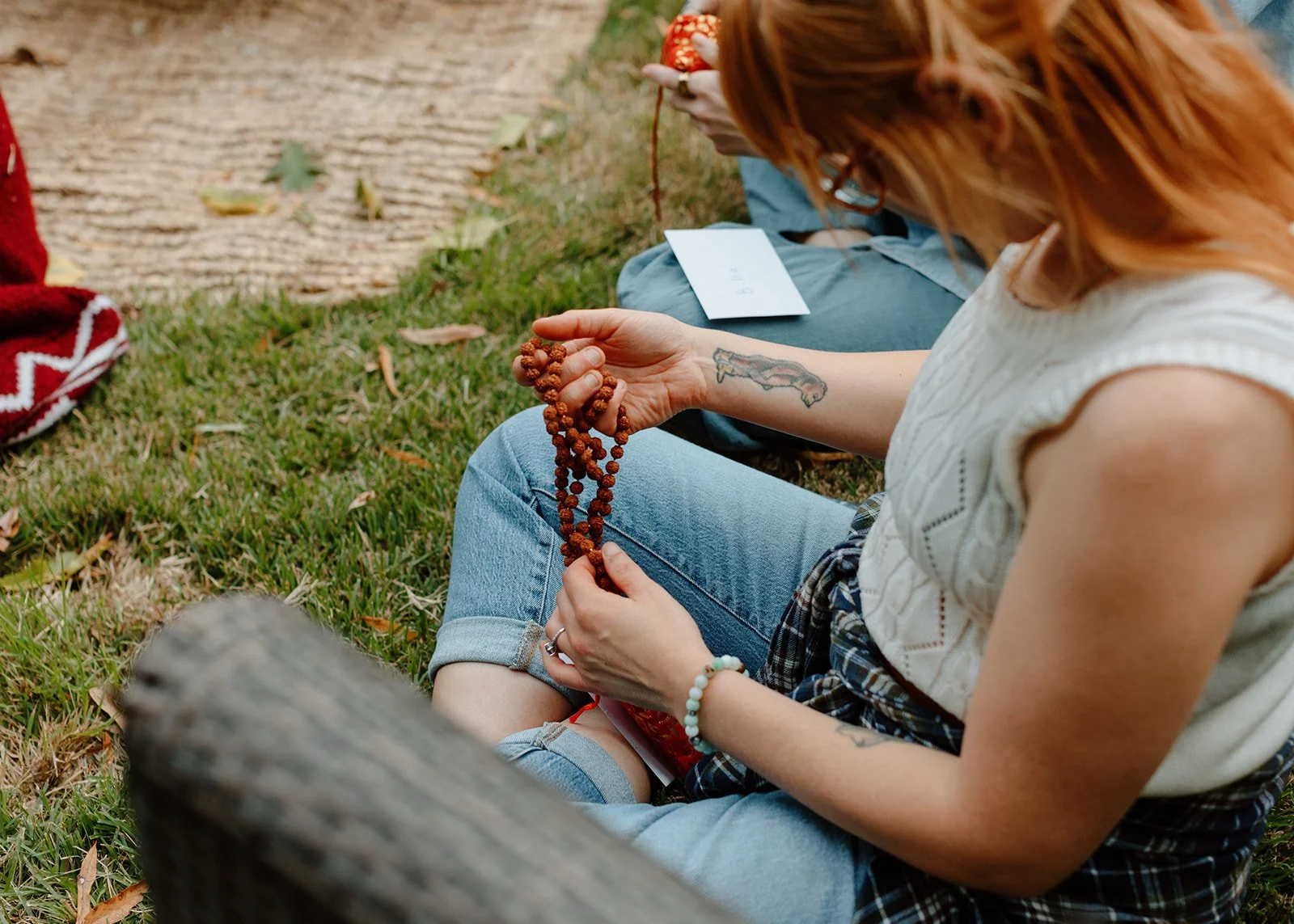 A person with red hair and a tattoo on their right arm sitting on grass, holding a string of prayer beads.