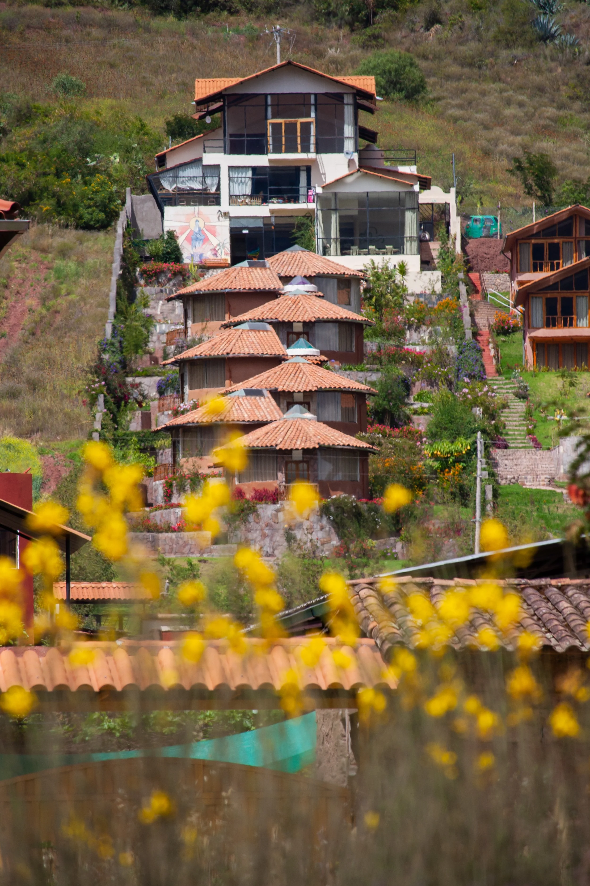 Seen from the perspective of the bottom of a hill and looking up, there are 7-circular buildings that are scattered back and forth up the hill with a larger white building at the top with lots of windows. Yellow flowers are blurred in the foreground.