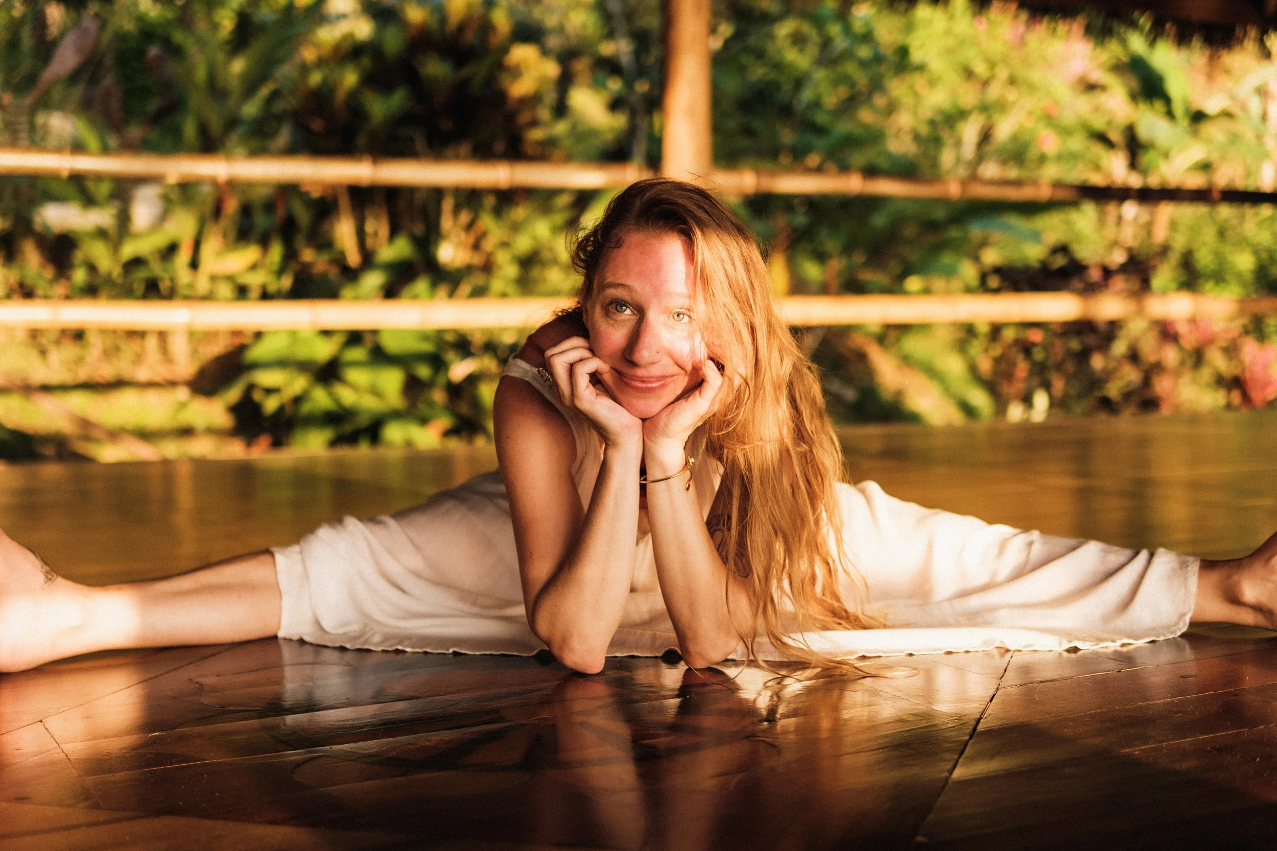 A blonde-haired person doing a full split on a wooden floor outdoors smiling and resting the chin on hands, with a blurred green and yellow garden background.