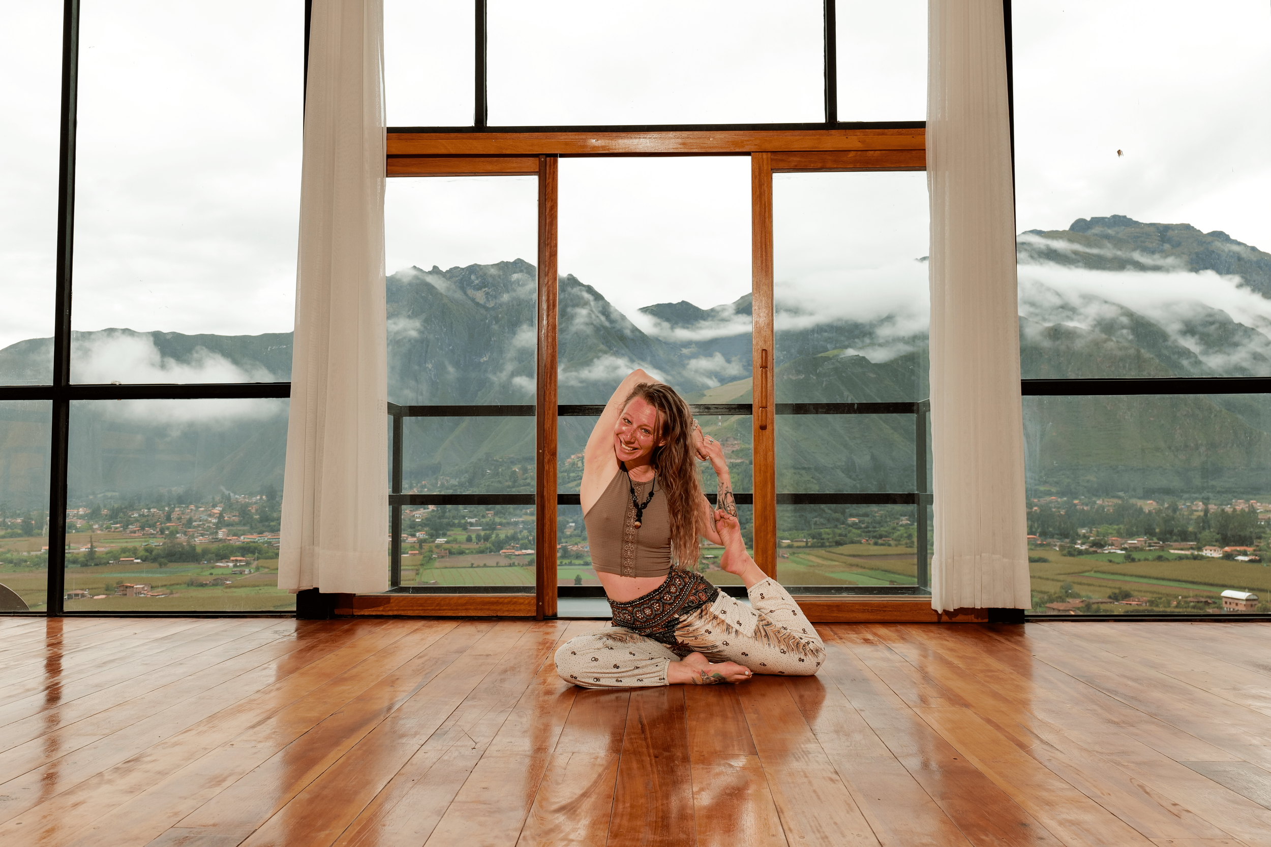 A blonde human is on the floor in an asymmetrical seat with her front leg bent in and her back leg bent bent up and held in place by her hands. She is smiling and there are foggy mountains in the background.