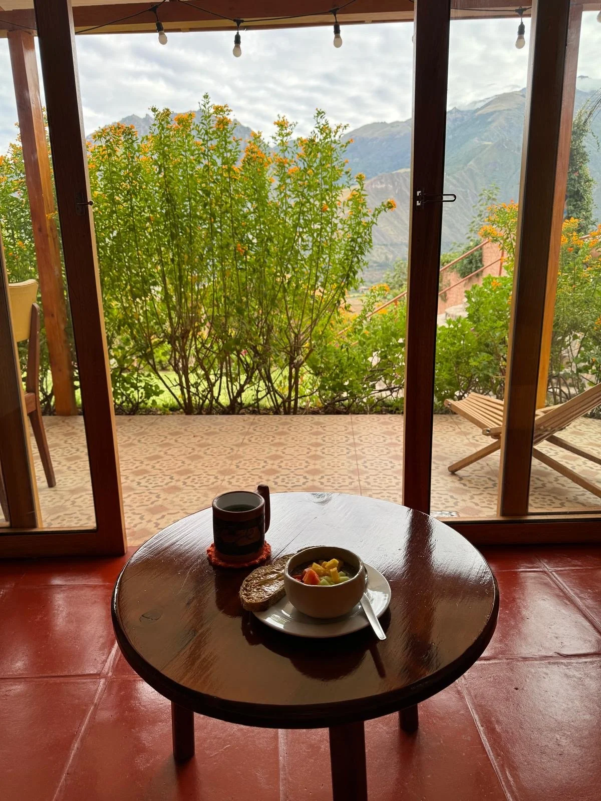 Coffee, bread and a fruit bowl sit on a small circular table before an open door with tall green plants as an outdoor wall with mountains, clouds, and sky in the background.