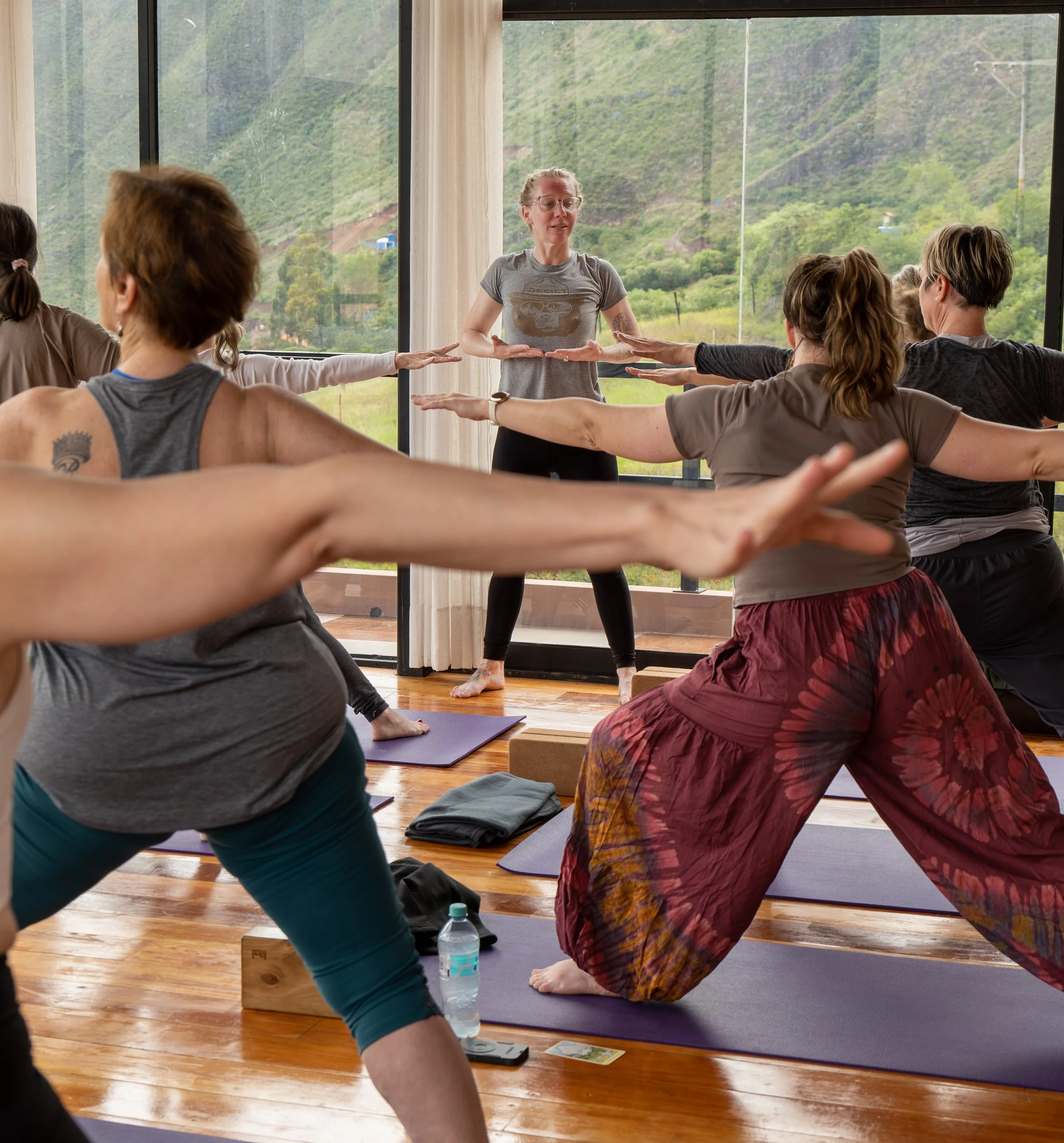 A group of people participating in a yoga class near large windows with a view of green hills and trees. They are doing a stretching pose with arms extended and are on yoga mats.