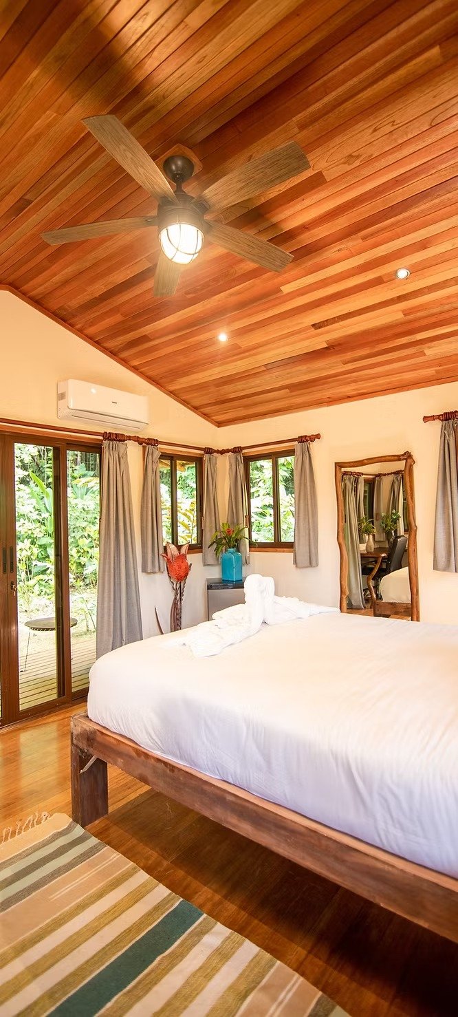 A cozy bedroom with a wooden ceiling fan, beige curtains, and a large window showing greenery outside. The room has a white bed with towel art, a mirror, and a river-blue vase with plants on a bedside table.