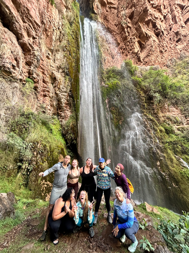 A group of 8 people huddle together with big smiles (and some tongues out) in front of a large waterfall amongst red cliffs and green trees.