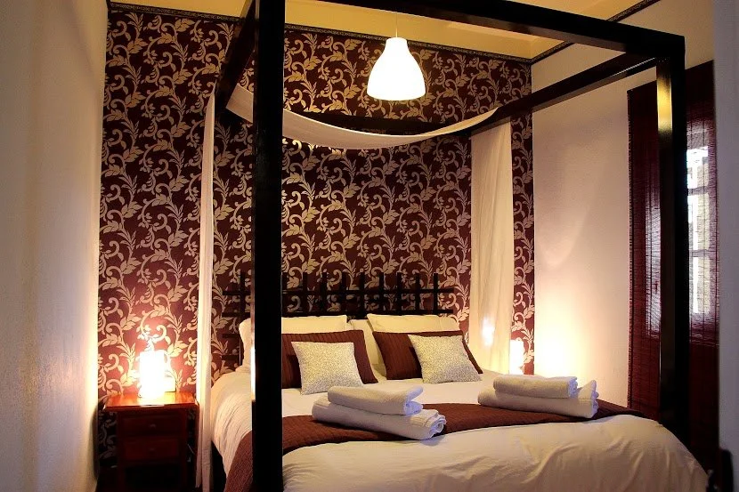 A bedroom with a four-poster bed, patterned wallpaper, bedside table with lamp, and white curtains.