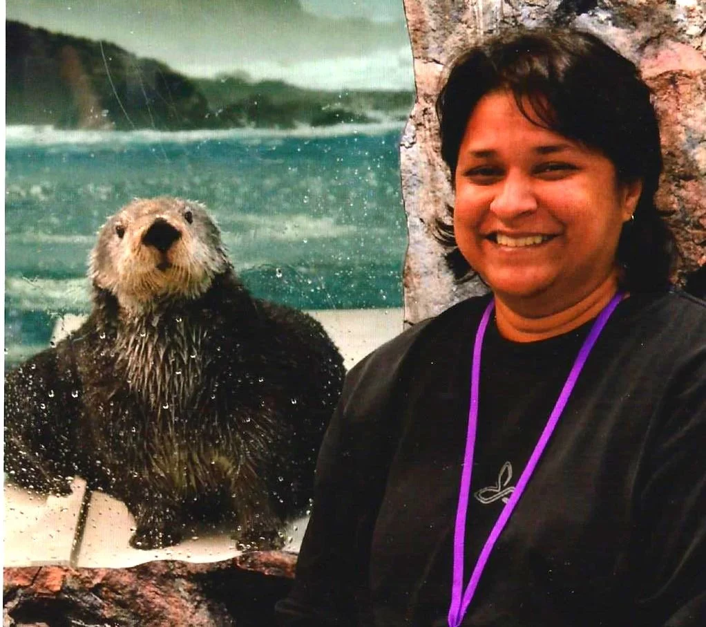 A person with short dark hair smiling, standing next to a wall that resembles a rocky surface, with a large poster of a sea otter in water behind her.