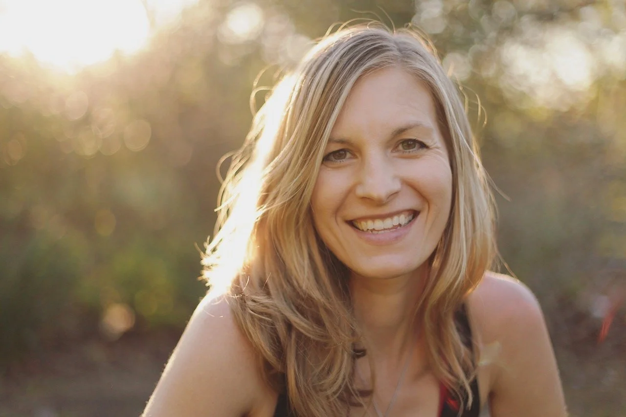 A person with long blonder hair smiling outdoors during sunset.