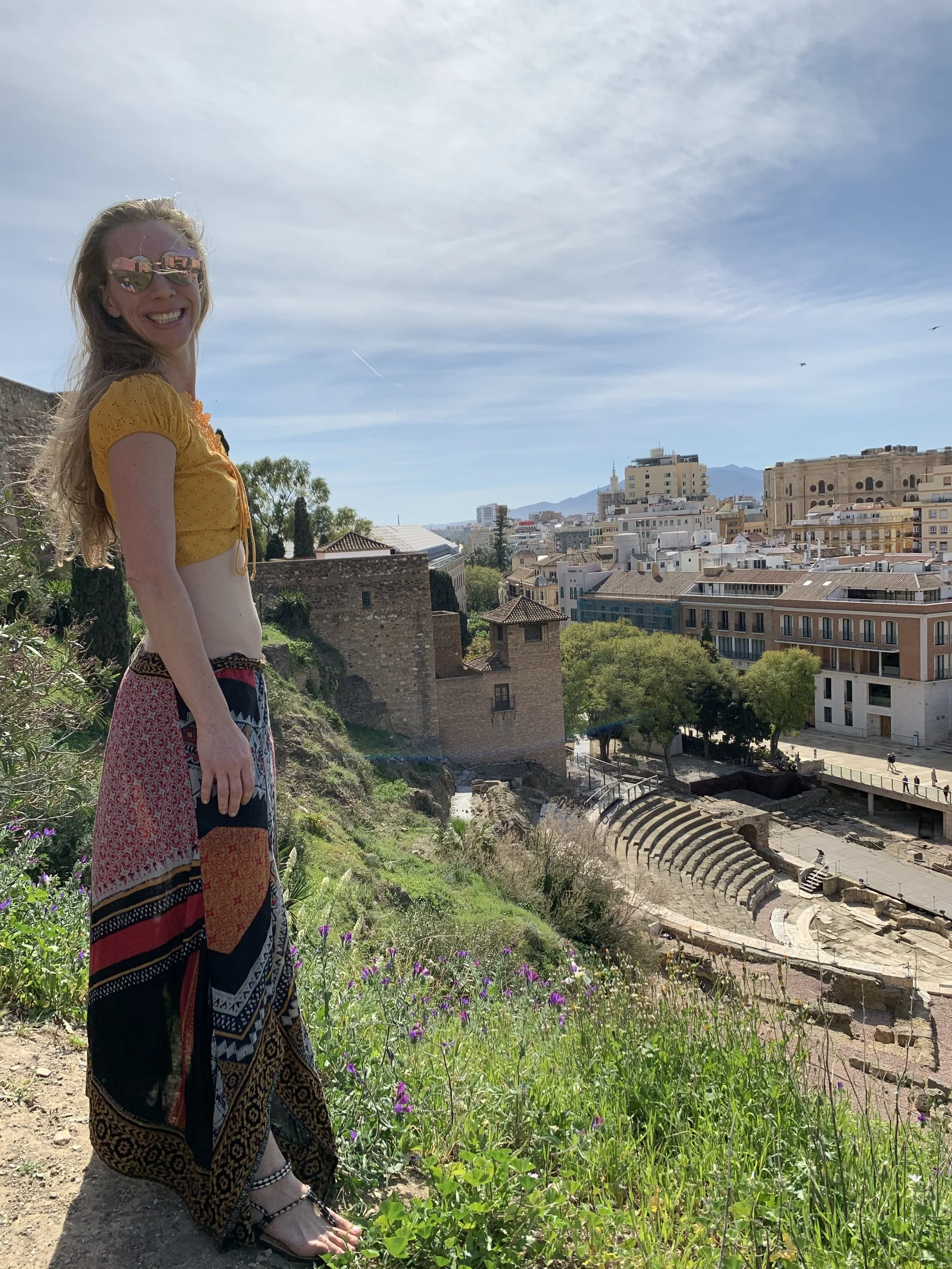 A smiling person in sunglasses and colorful clothing stands beside an ancient outdoor theater with a hillside cityscape, mountains, and blue sky in the background.