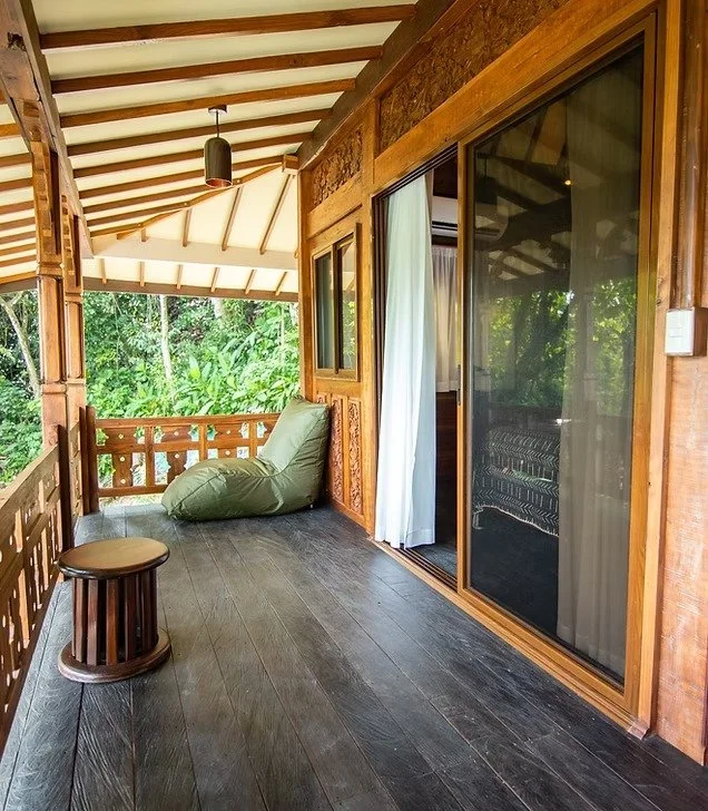 Wooden balcony with sliding glass door, curtain, and green bean bag chair overlooking greenery.