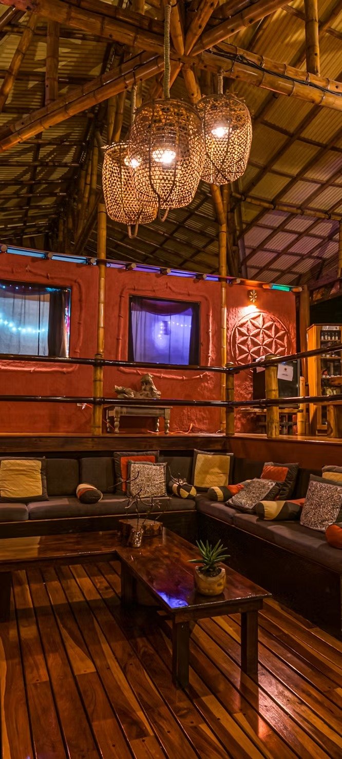 Interior of a cozy lounge area with bamboo and wood decor, hanging wicker lanterns, a sofa with cushions, a wood coffee table, and illuminated windows and walls.