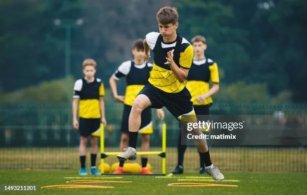 A young soccer player jumping over agility hurdles during training with teammates in the background on a field.