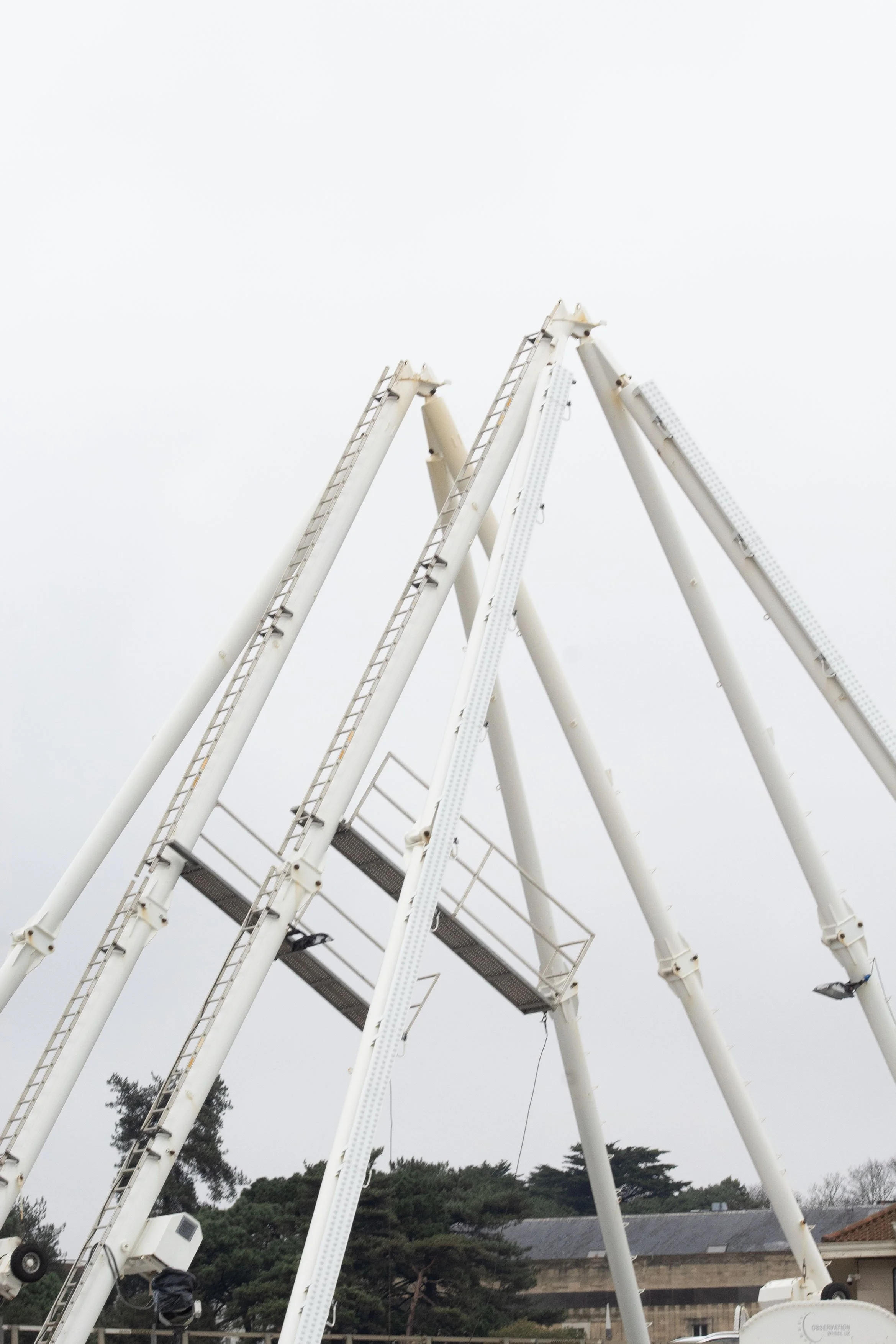A close-up view of white amusement park ride arms with ladders, set against a cloudy sky. Trees and a building are visible in the background.