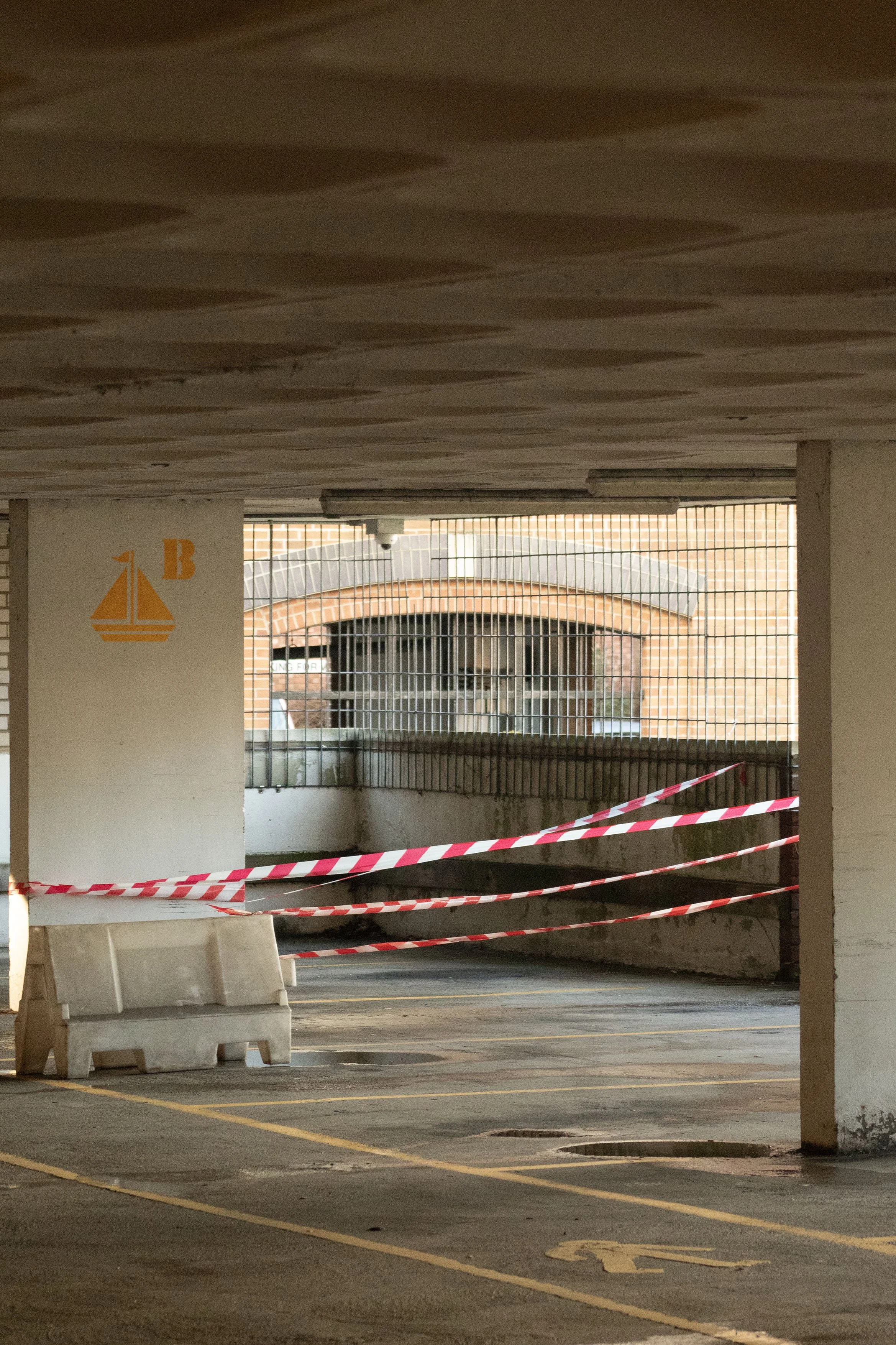 Underground parking garage with concrete columns, security camera, and a brick building visible through a barred window, with red and white tape blocking part of the area.