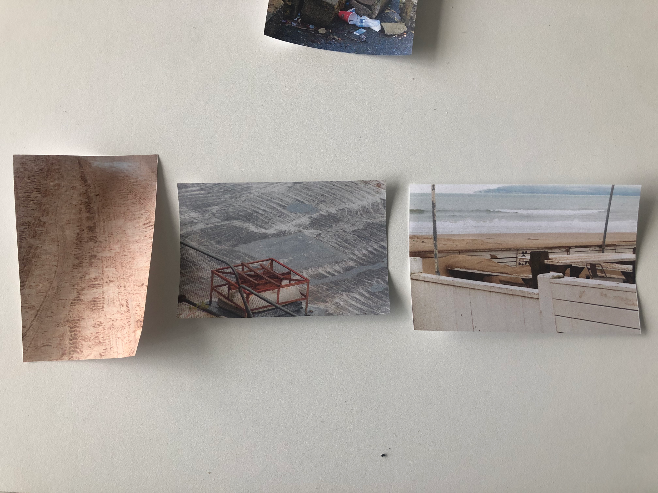 Several photographs are tacked onto a plain white wall. One photo shows a beach scene with waves and sandy shore. Another photo appears to depict a construction site with machinery and dirt. A third photo shows trash and rocks along a rough ground. T